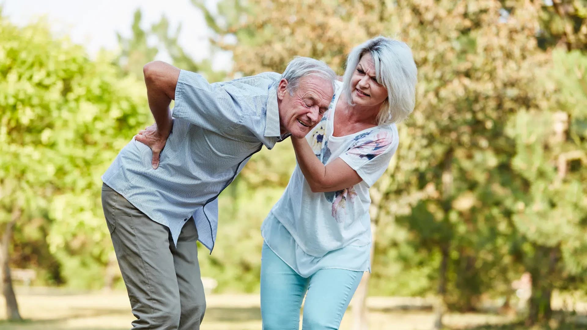 Older man bending forward holding his lower back as an older woman supports him outdoors
