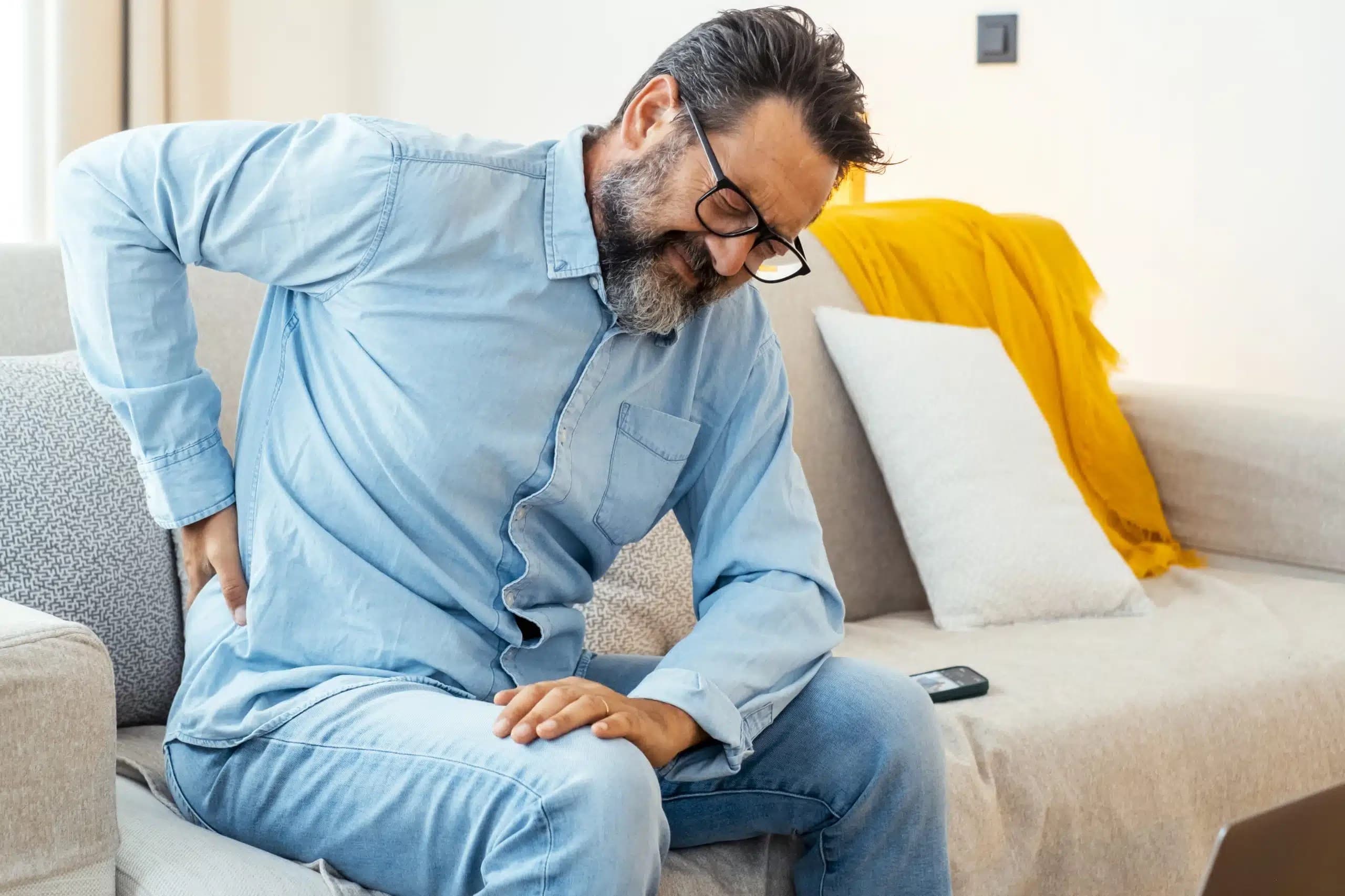 Bearded man with glasses sitting on a sofa, holding his lower back in pain