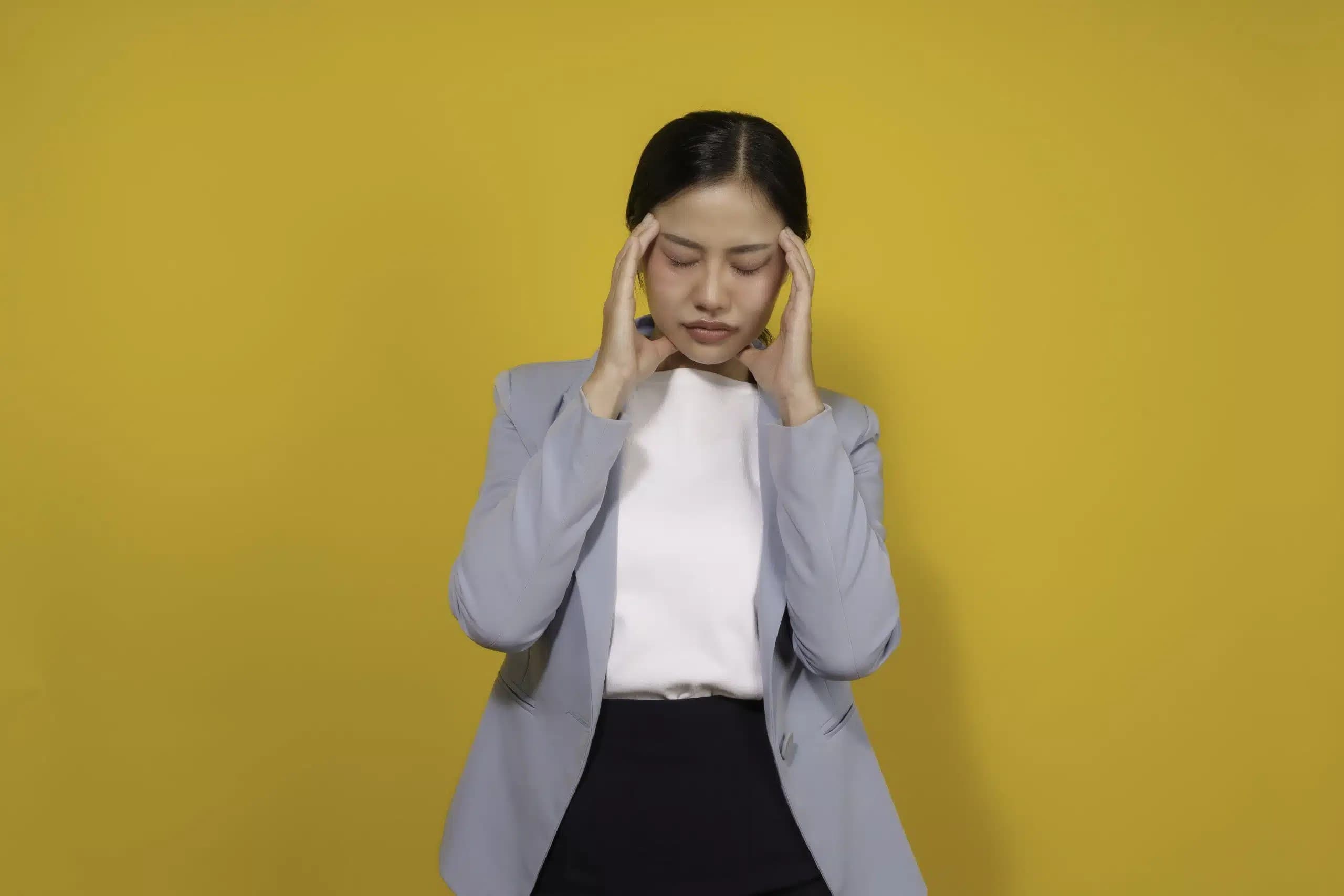 Woman with her eyes closed pressing both temples against a yellow background