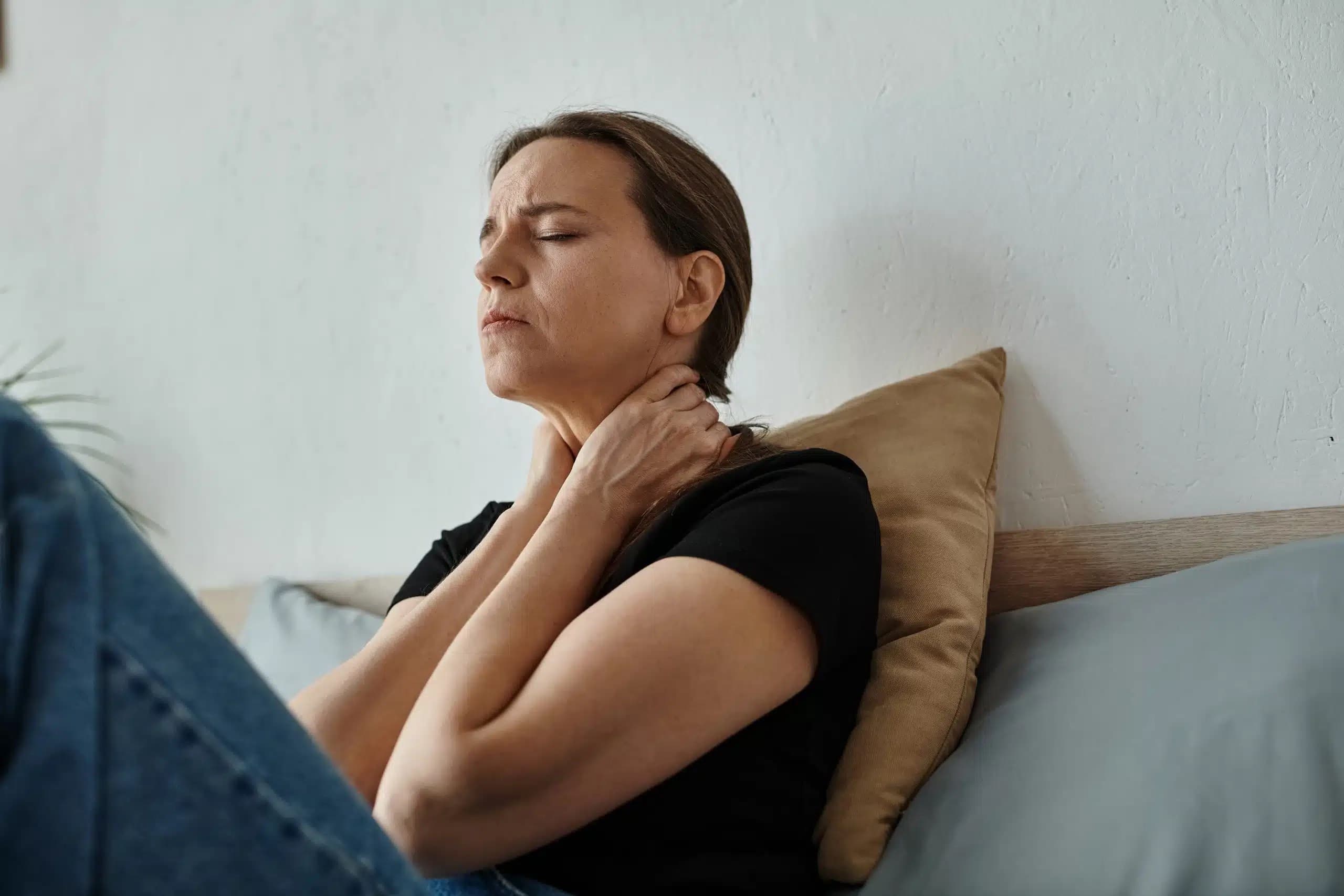 Woman sitting against pillows with her eyes closed, holding her neck in pain