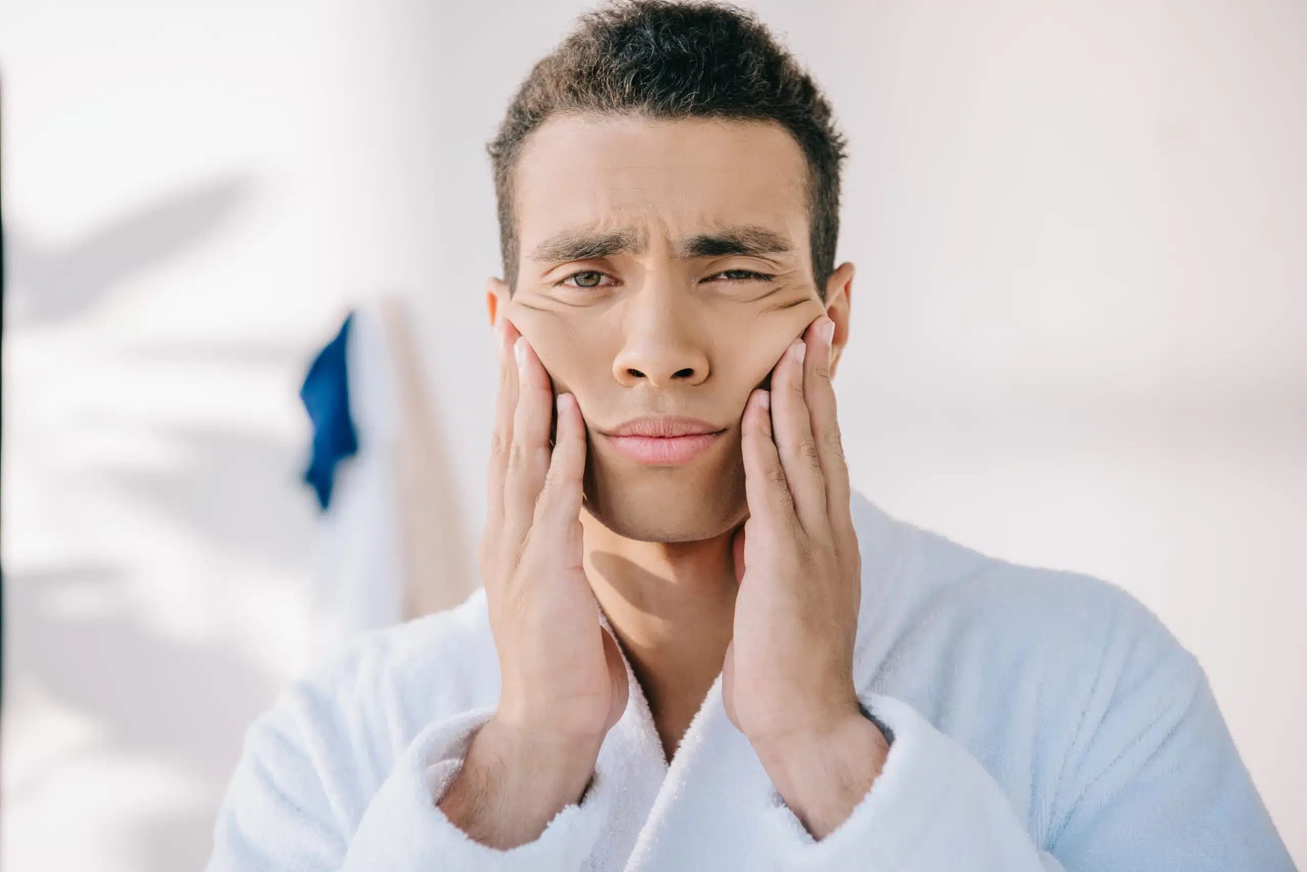 Man in a white bathrobe pressing both hands against his cheeks with a pained look