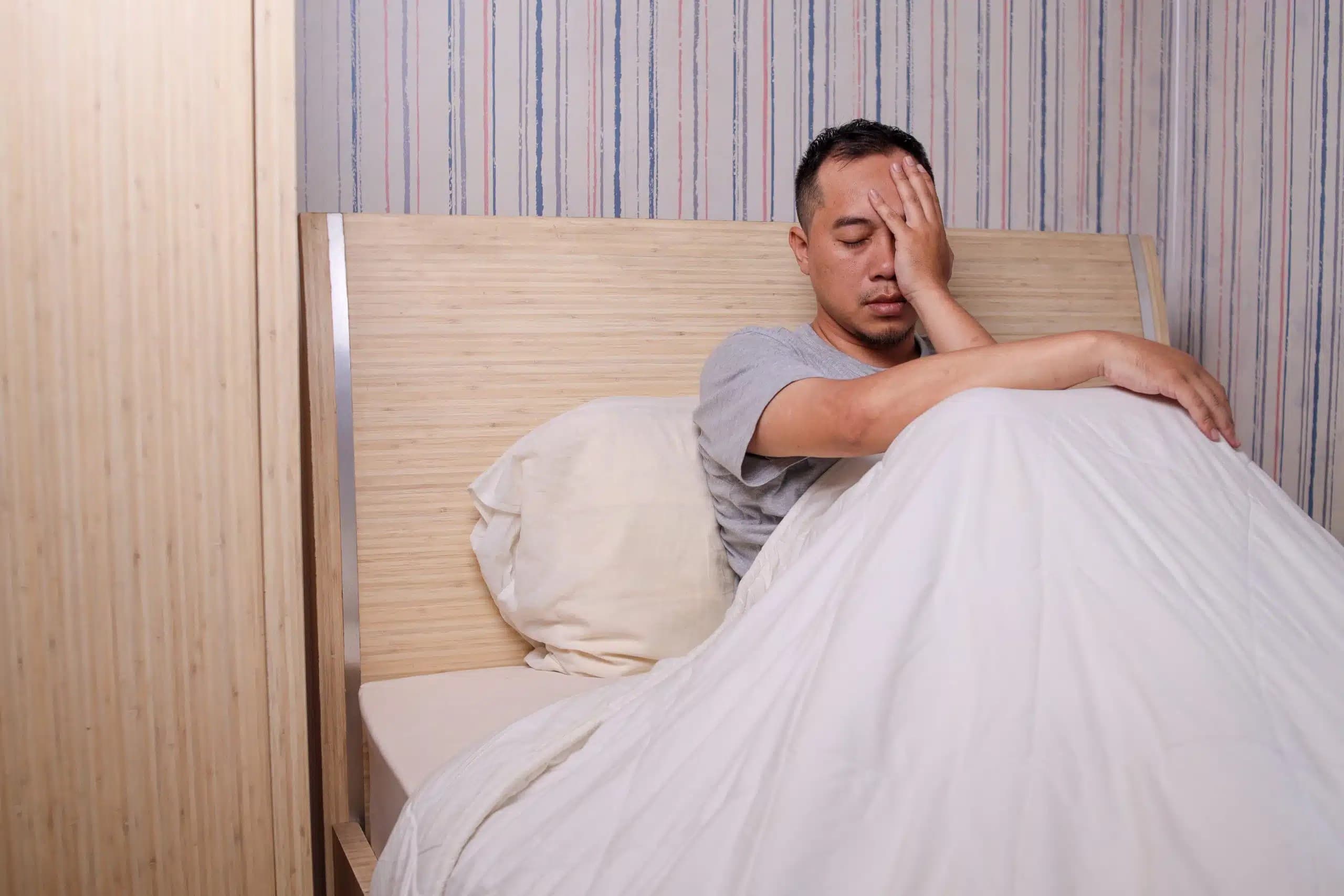 Man sitting up in bed under a white duvet, covering his eyes with one hand