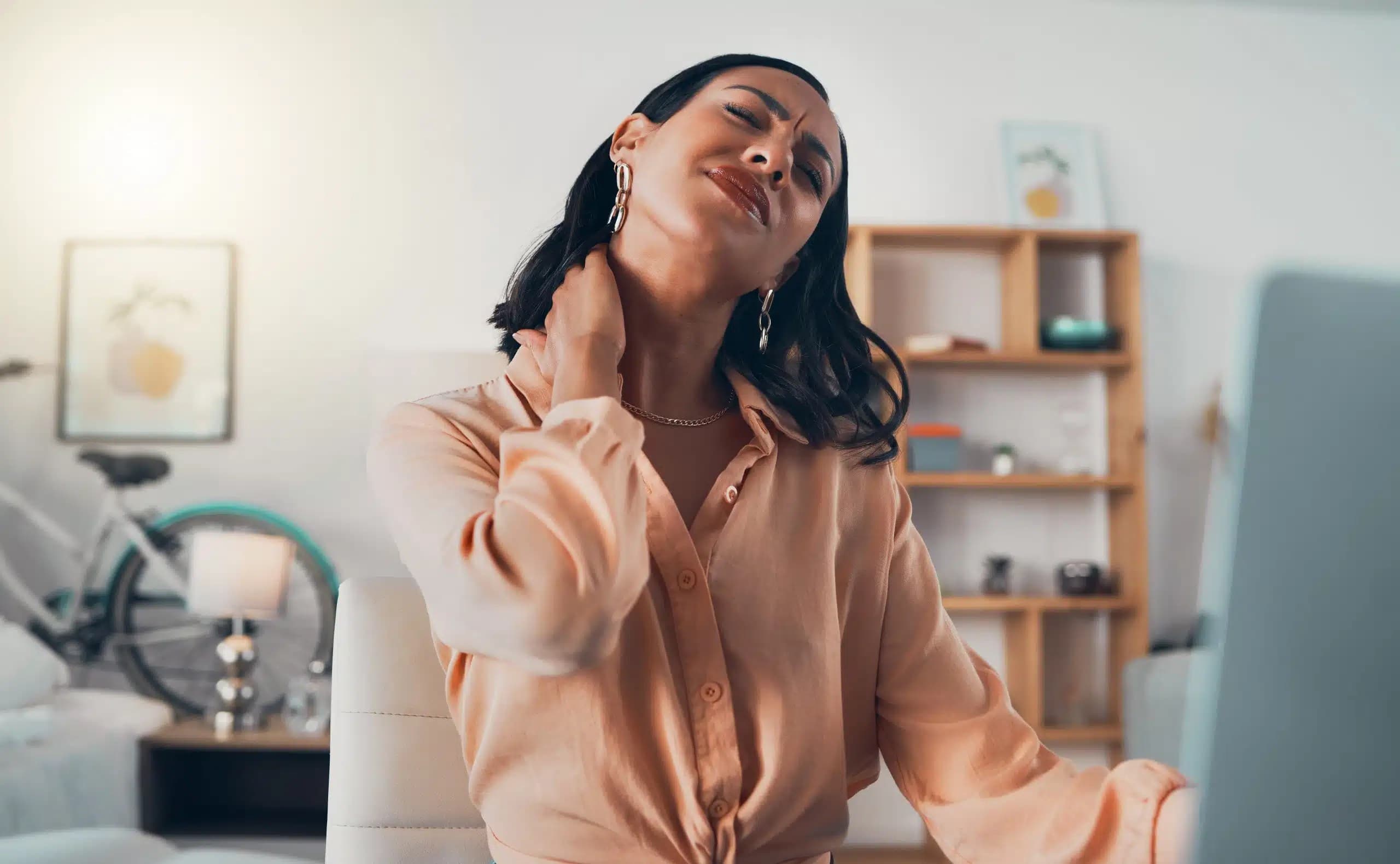 Woman in a bright home office tilting her head and rubbing the back of her neck, wincing