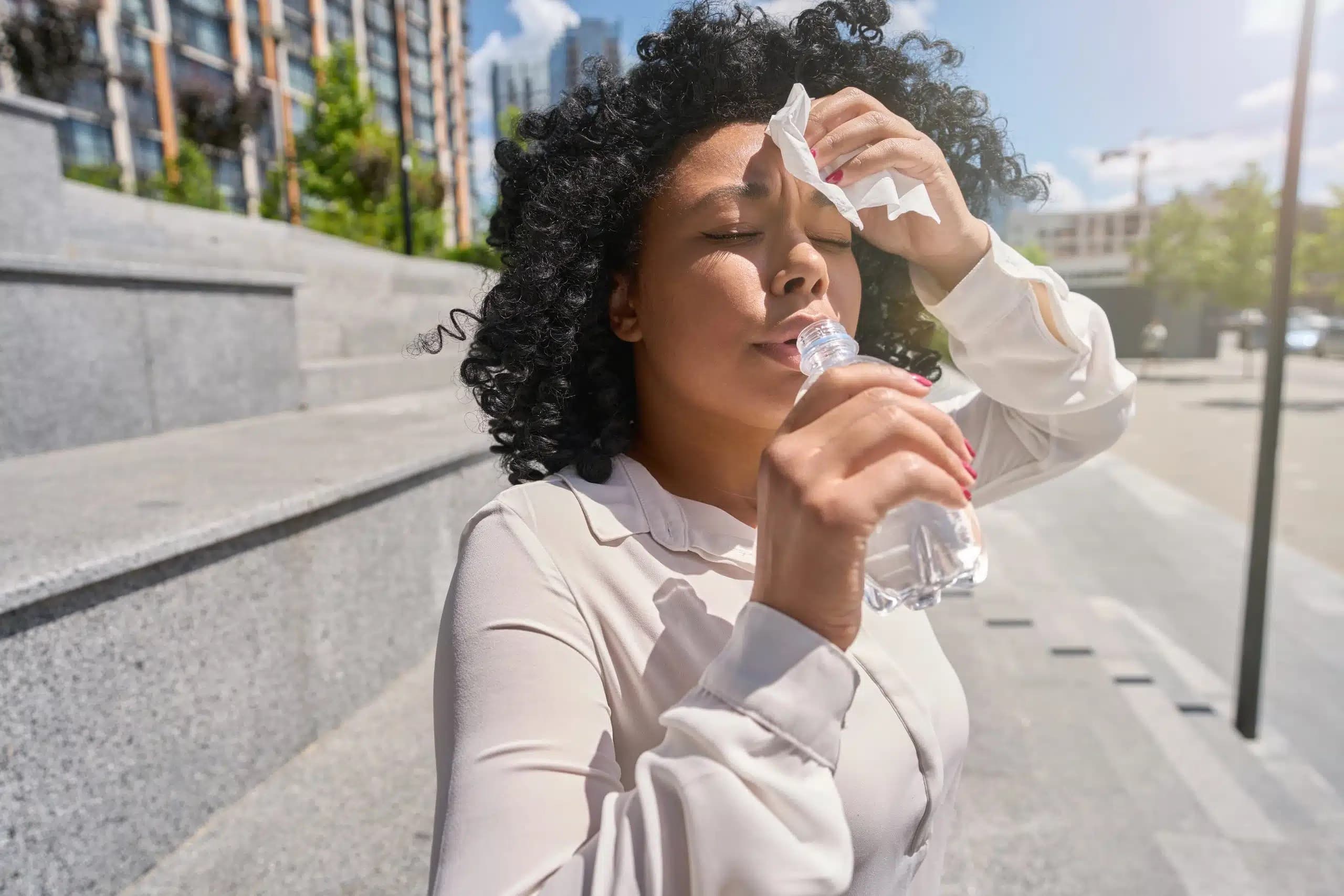Woman outdoors holding a plastic water bottle and a tissue to her forehead in the heat