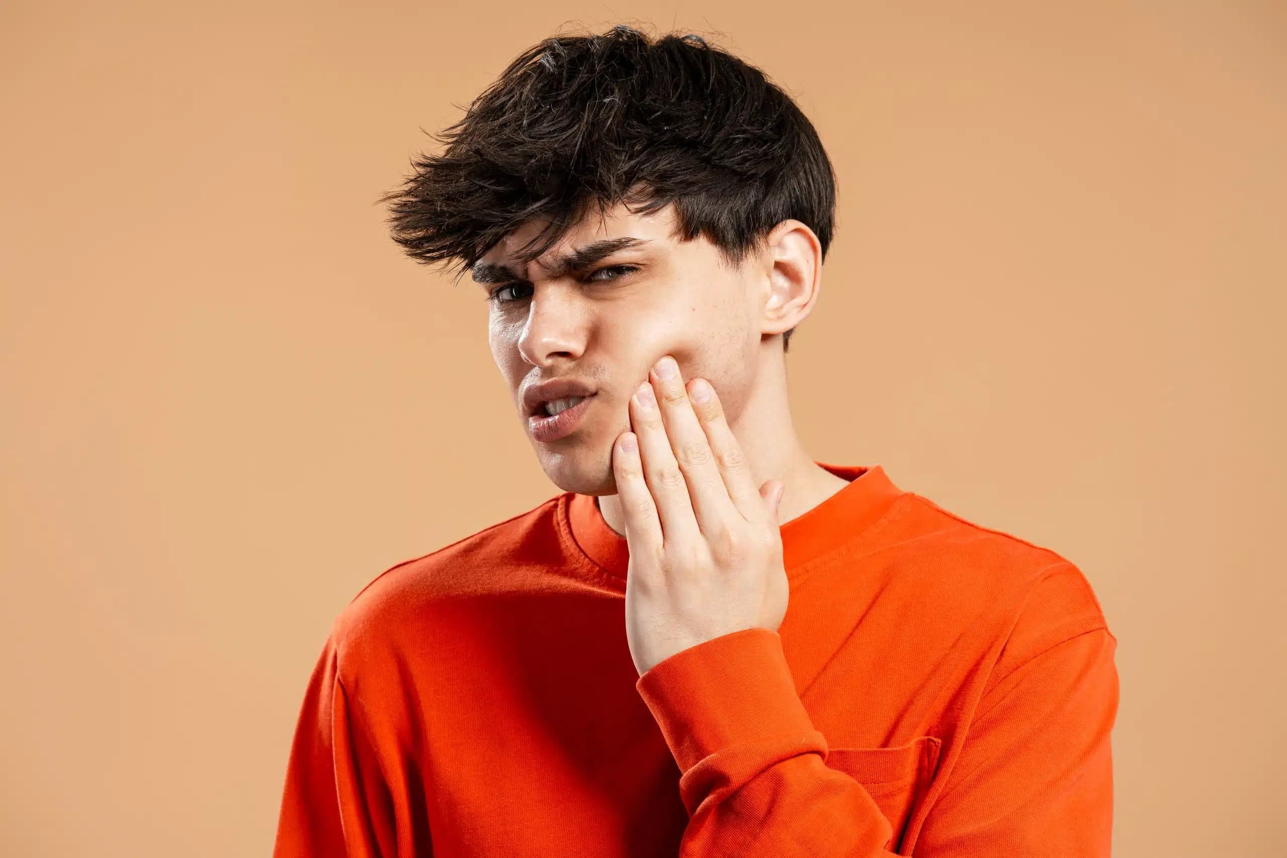 Young man in an orange sweater wincing with his hand against his cheek