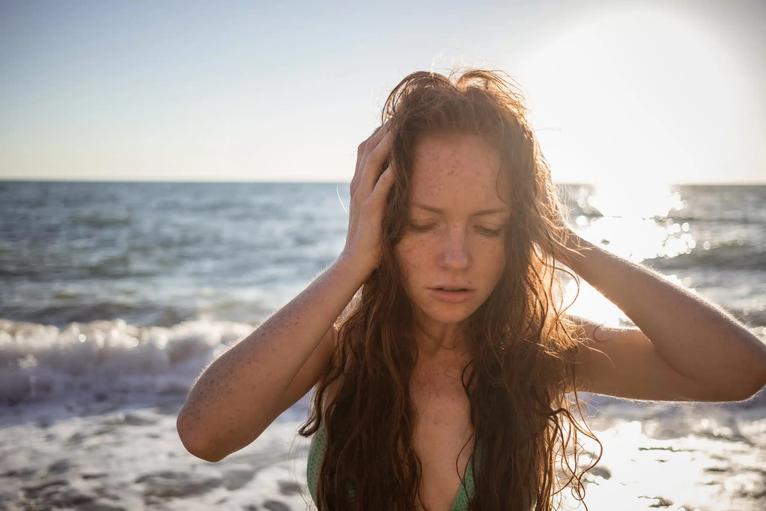 Woman with wet hair on a sunlit beach, holding her head with both hands and eyes closed