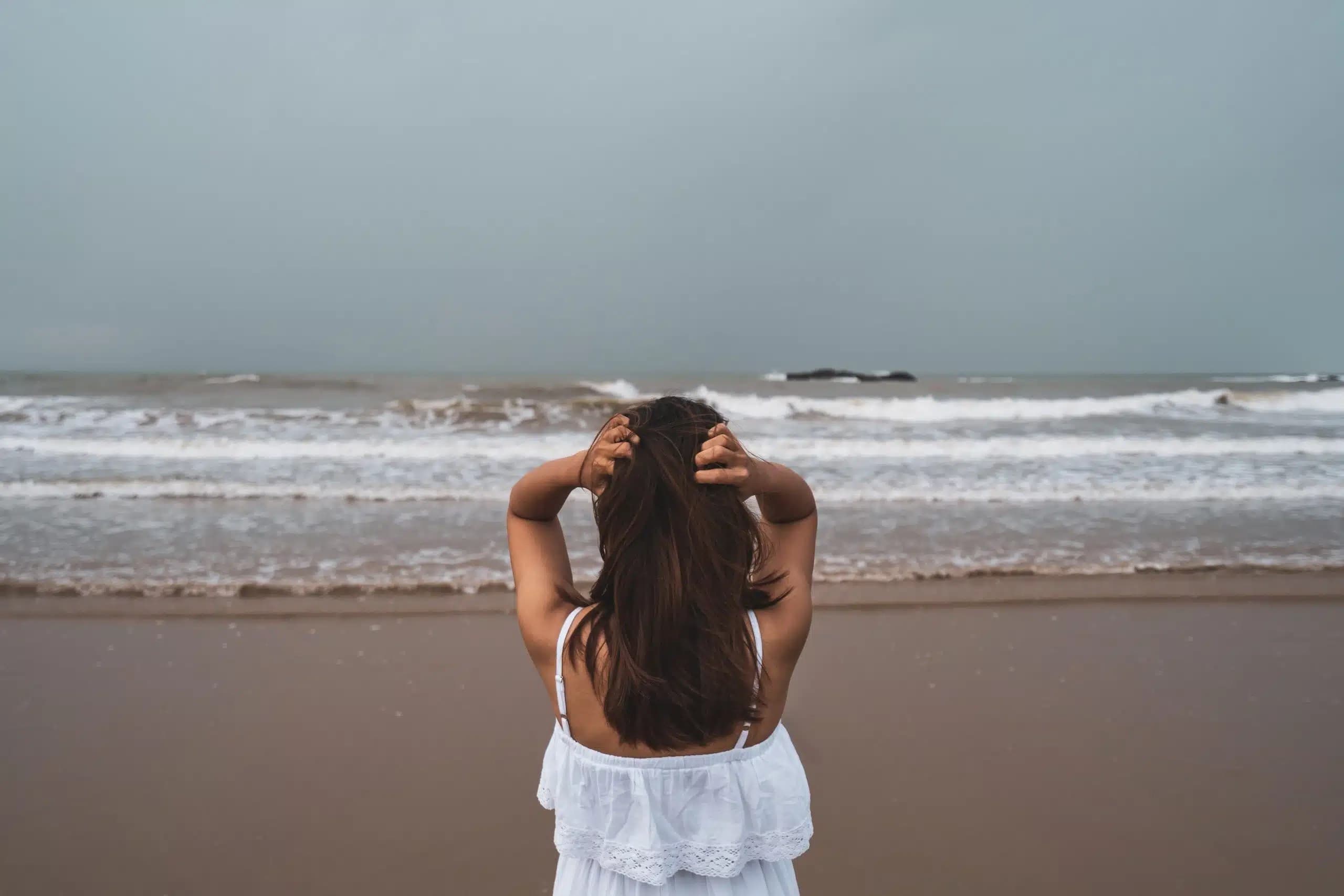 Woman seen from behind on a cloudy beach, holding her head with both hands