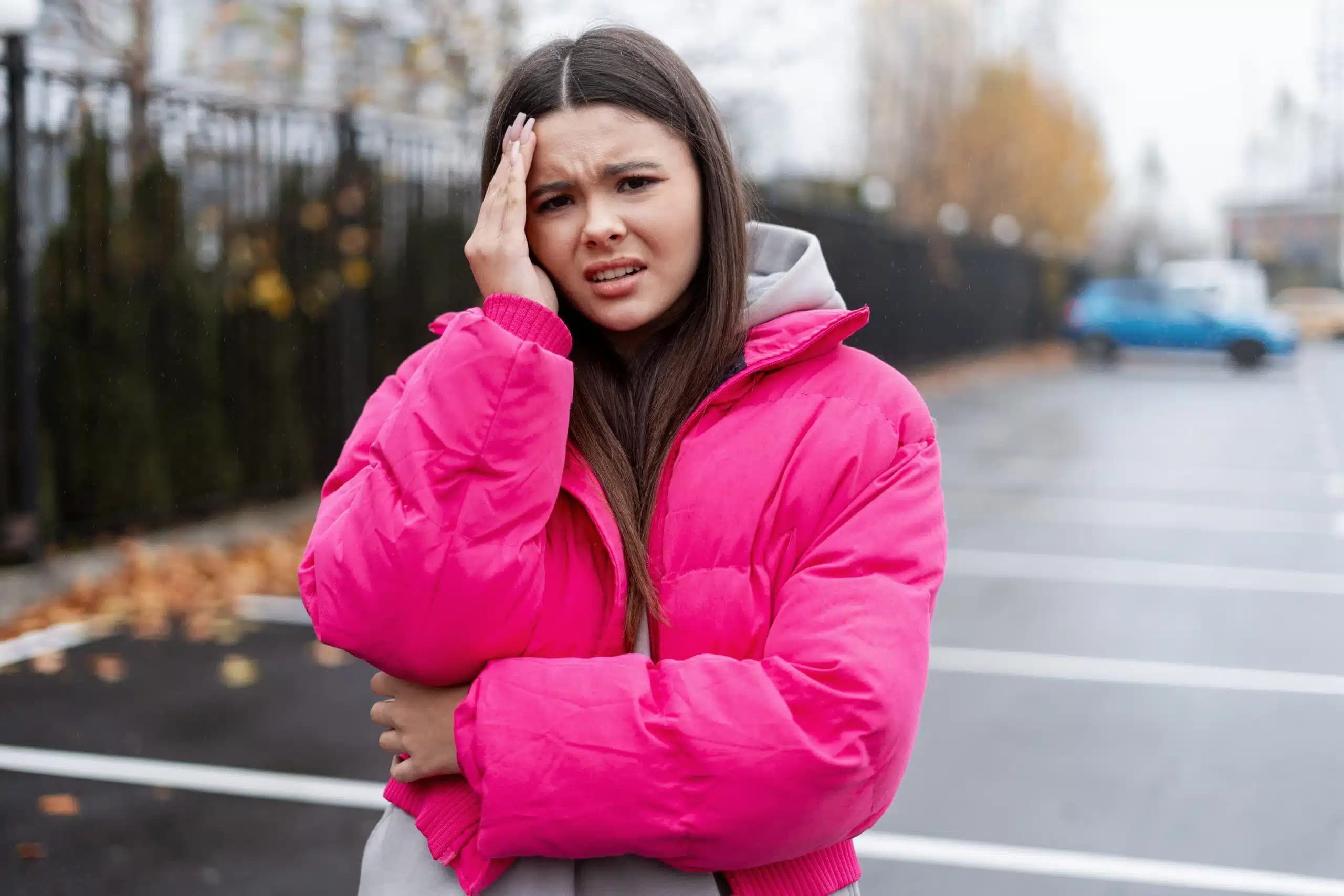 Woman in a pink jacket outdoors with her hand on her forehead and a pained expression