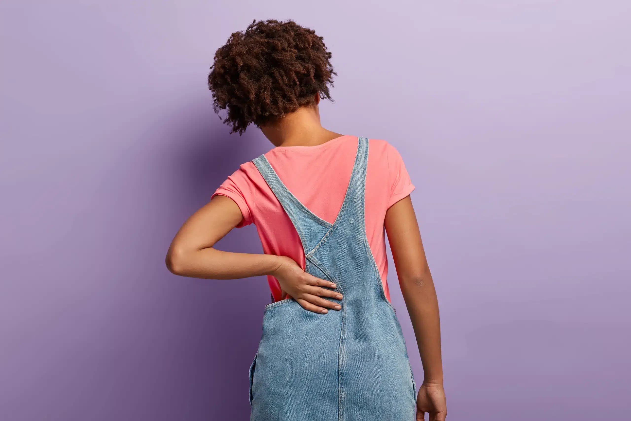 Woman seen from behind against a purple wall, holding her lower back